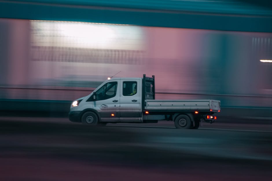 A silver flatbed van operated by Officeremovals.org.uk is moving along a city street at dusk or nighttime with blurred brightly lit buildings in the background, creating a sense of motion. The van is