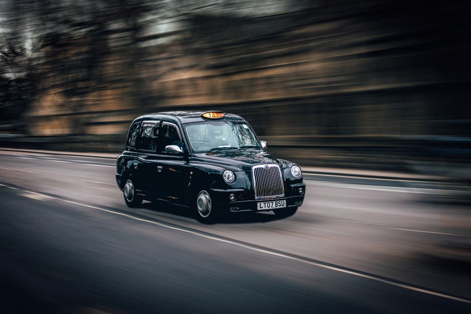 A silver flatbed van operated by Officeremovals.org.uk is moving along a city street at dusk or nighttime with blurred brightly lit buildings in the background, creating a sense of motion. The van is