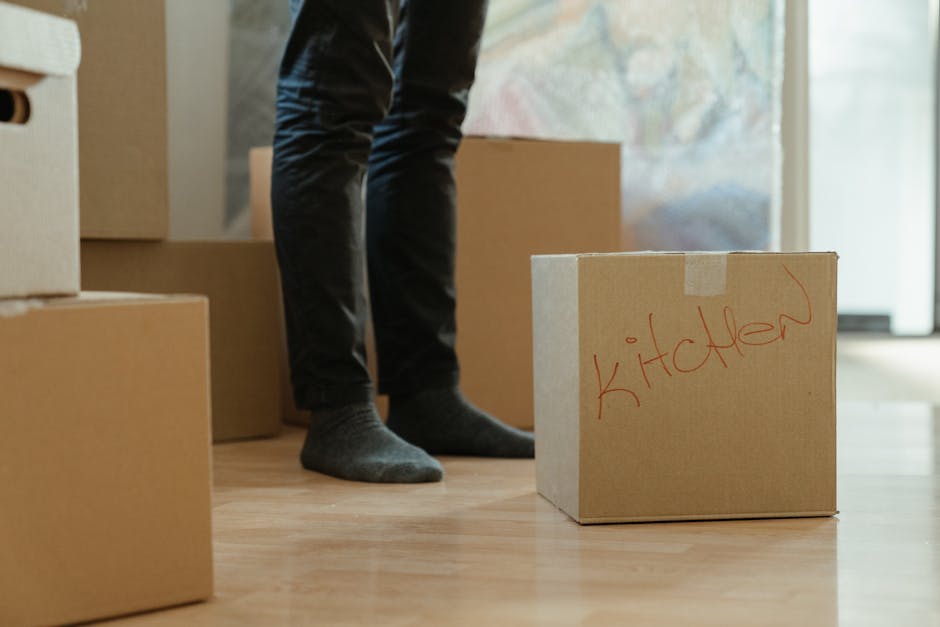Close-up of a person's hand placing bubble wrap inside a cardboard box labeled 'KITCHEN' during a home relocation process, with the box partially open and positioned on a flat surface inside a propert