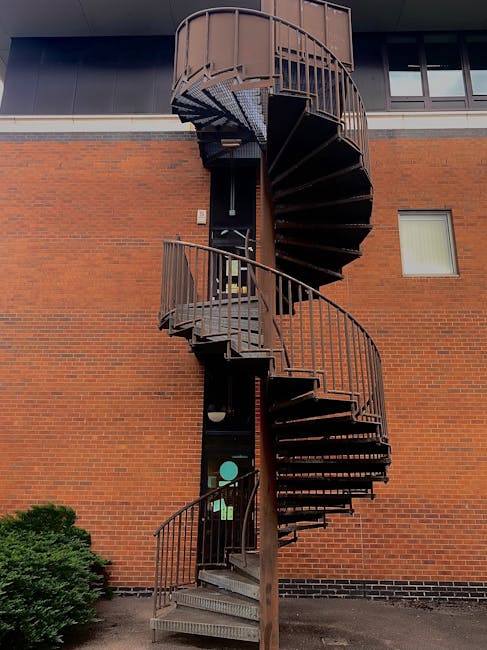 A tall, external metal spiral staircase with brown railings and dark steps is attached to the brick exterior wall of a building. The staircase curves upward around a central support pole, leading to a